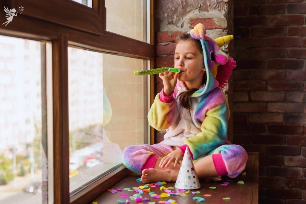 girl ina unicorn onsie sitting alone in a window blowing a party whitle