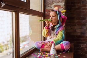 girl ina unicorn onsie sitting alone in a window blowing a party whitle