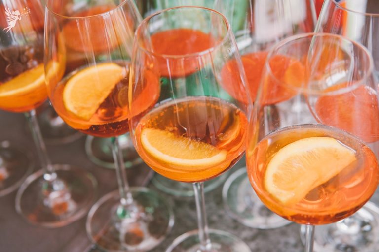 close up of 4 glasses aperol spritz being prepared with orange slice garnishes added