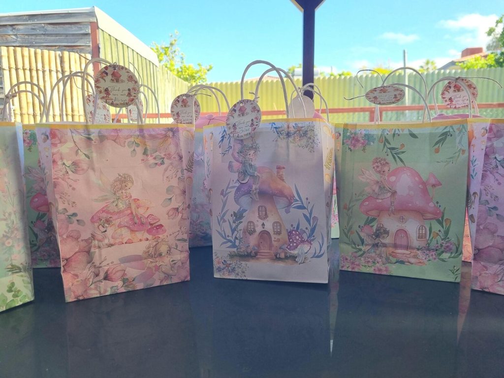 A collection of pre-packed watercolor fairy and mushroom house illustrated gift bags with personalized round thank you tags and rope handles displayed outdoors make beautiful ready to go fairy party favors for a girls birthday celebration