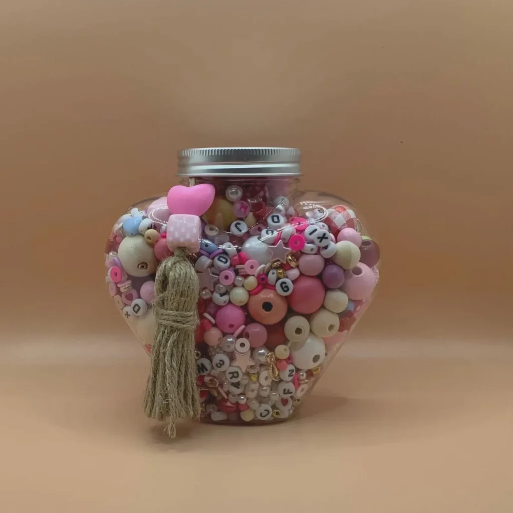 Heart-shaped jar overflowing with pink, white, and wooden beads, letter beads, and charms for bracelet making — hands-on and charming tween girl party favors.