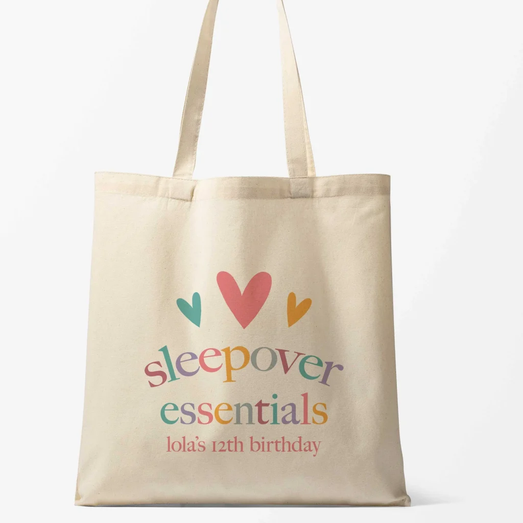 Natural canvas tote bag printed with colorful hearts and "Sleepover Essentials" text personalized for Lola's 12th birthday — practical and cute tween girl party favors.