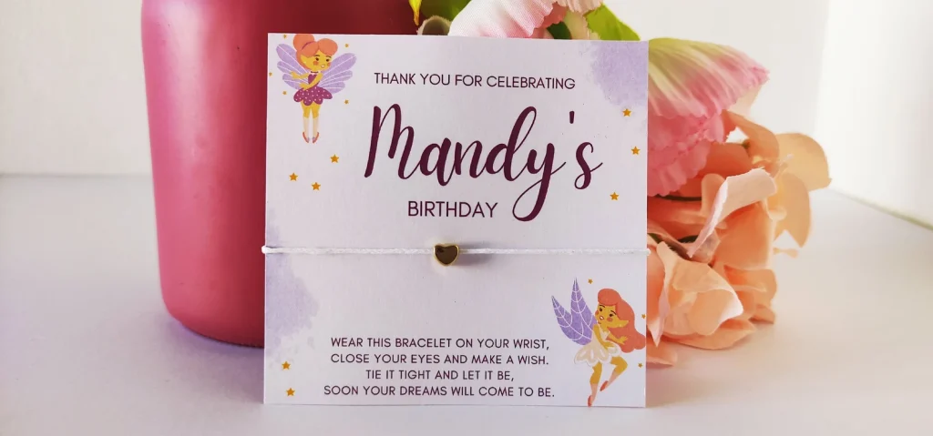 A delicate string wish bracelet with a gold heart charm displayed on a personalized purple watercolor fairy illustrated birthday card makes a heartfelt fairy party favor for kids