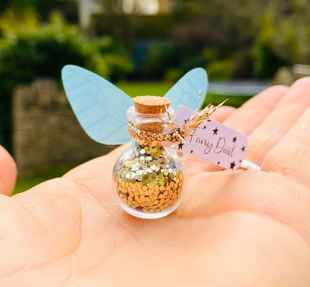 A palm sized round glass bottle filled with golden glitter decorated with teal fairy wings and a rose gold twine accent serves as a magical fairy party favor