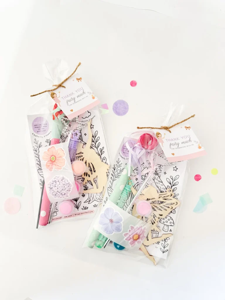 Two clear cellophane goodie bags filled with a wooden fairy cutout pastel paints flower stickers and a charm necklace tied with twine make creative fairy party favors for a girls birthday celebration