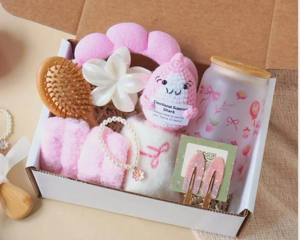 Pink gift box filled with a wooden hairbrush, fluffy socks, pearl bracelet, crochet shark plushie, glass cup, and hair clips — adorable tween girl party favors.