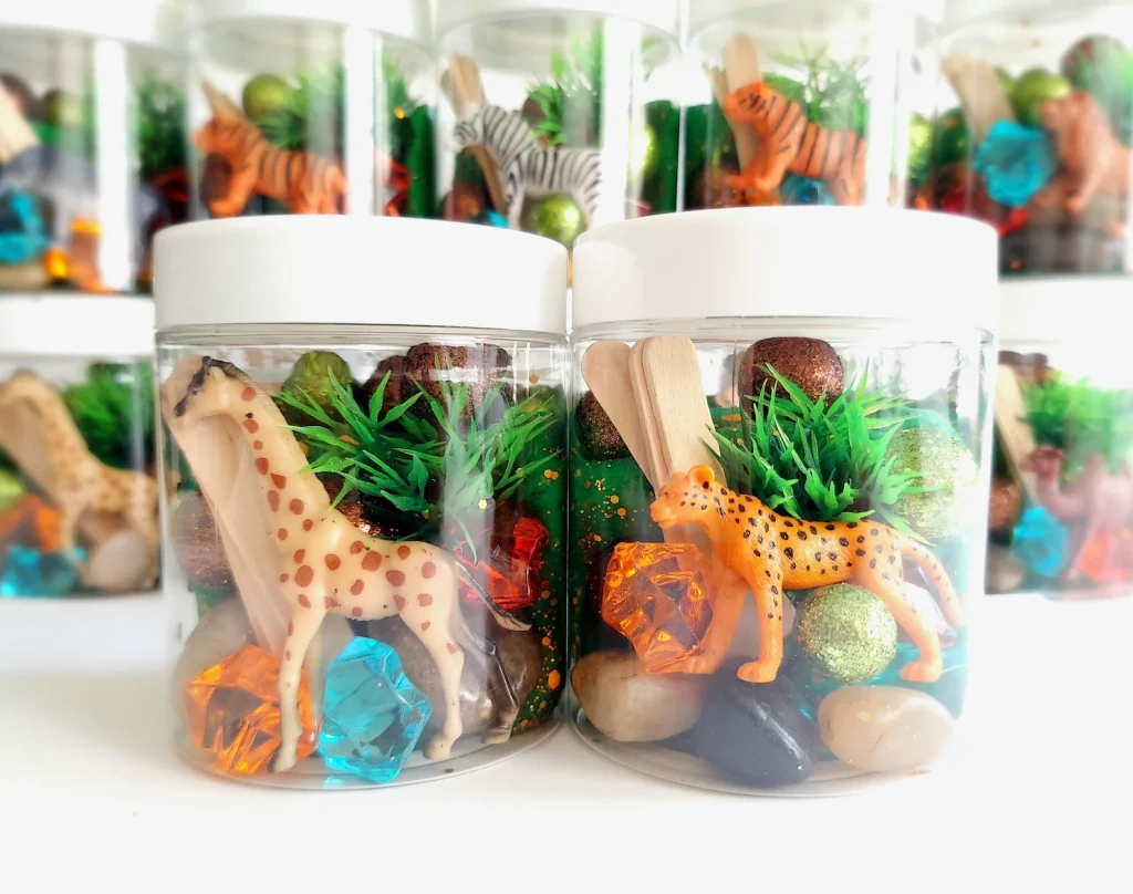 Safari animals party favor ideas featuring clear sensory playdough jars filled with jungle animal figurines, gemstones, pebbles, and green grass accents.