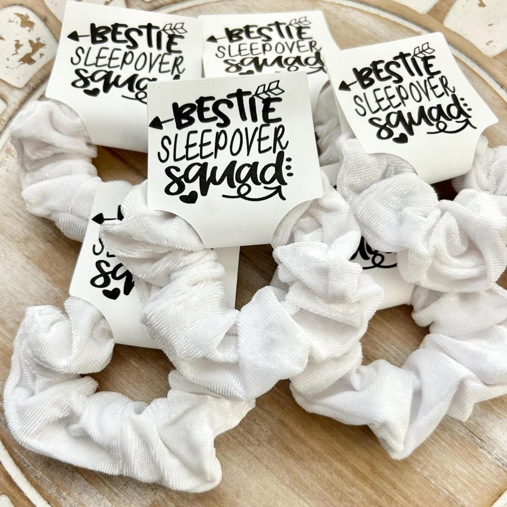 White velvet scrunchies on "Bestie Sleepover Squad" cards displayed on a rustic wooden tray, serving as fun and fashionable tween girl party favors.