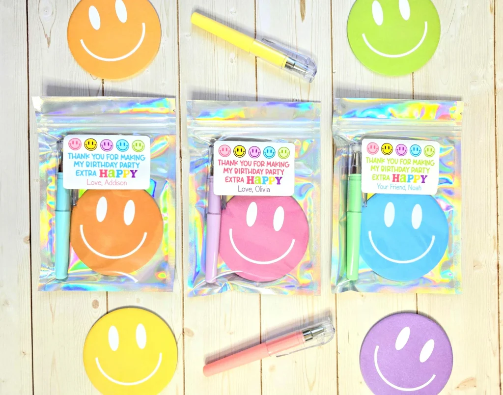 Holographic bags containing colorful smiley face notepads and pens with personalized "Thank You for Making My Birthday Party Extra Happy" tags — cheerful tween girl party favors.