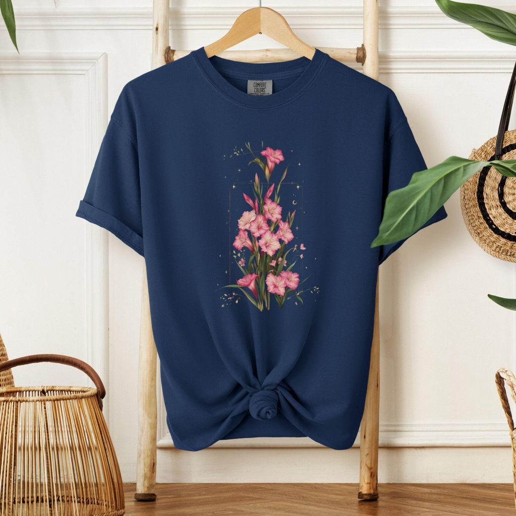 A navy cotton tee hanging on a ladder featuring a celestial pink gladiolus stem illustration with stars and a moon, a wearable August birthday gift idea.