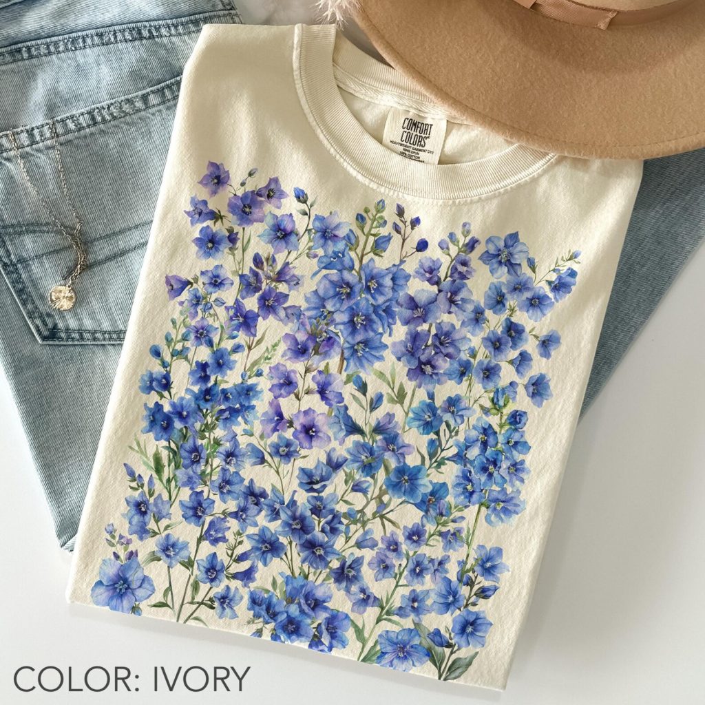 A folded ivory cotton tee printed with a dense watercolor cluster of blue and purple larkspur blooms, a wearable and charming July birthday gift idea.
