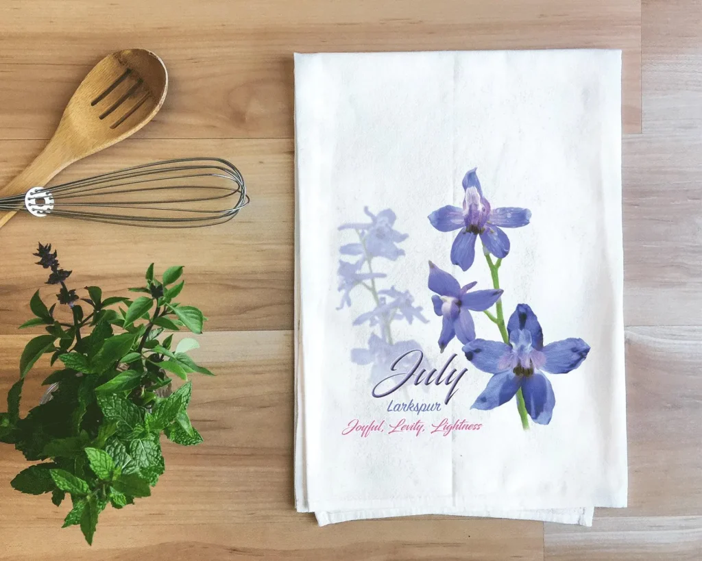 A white cotton tea towel printed with purple larkspur blooms and the birth month flower meaning, a sweet and practical July birthday gift idea.