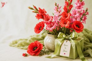A green gift box with a satin bow beside a ceramic vase of red poppies and pink gladiolus blooms, inspiring beautiful August birthday gift ideas.