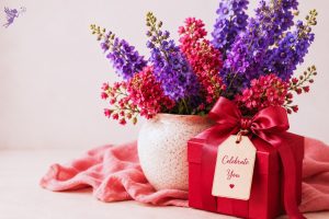 A red gift box with a satin bow and celebrate tag beside a ceramic vase of vibrant purple and pink larkspur blooms, inspiring elegant July birthday gift ideas.