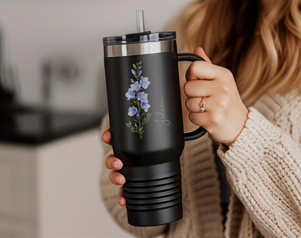 A matte black insulated tumbler with a handle featuring a blue larkspur stem and a personalized name in script, a practical July birthday gift idea.