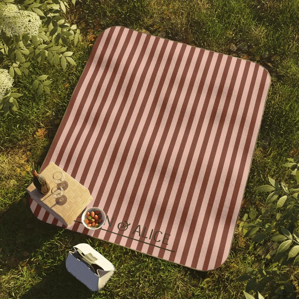 A personalized red and pink striped picnic blanket laid on grass beside a wicker basket and strawberries, a thoughtful and practical July birthday gift idea.