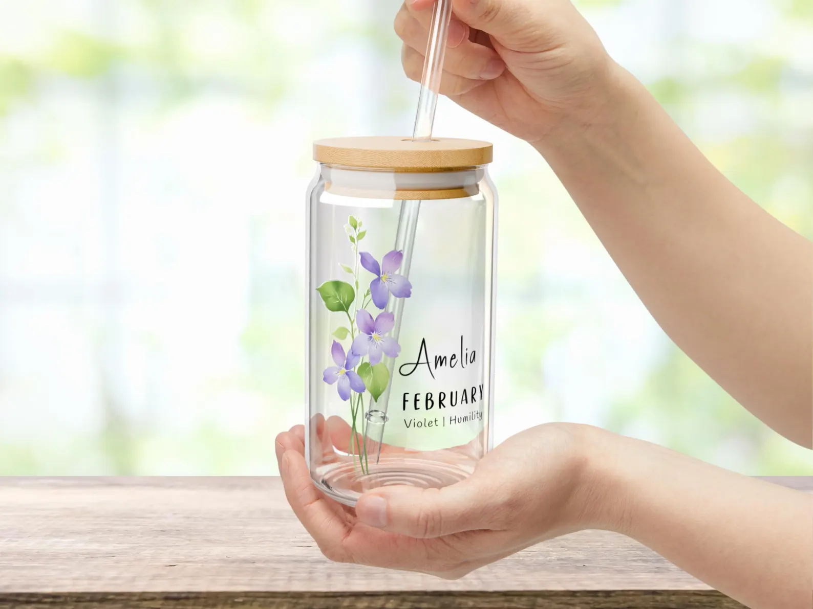 A glass can tumbler with a bamboo lid and glass straw printed with purple violet flowers and a personalized name, a pretty February birthday gift idea.