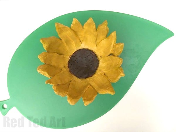 A golden clay bowl with a sculpted sunflower design on a green leaf background, a lovely homemade gift for grandparents.