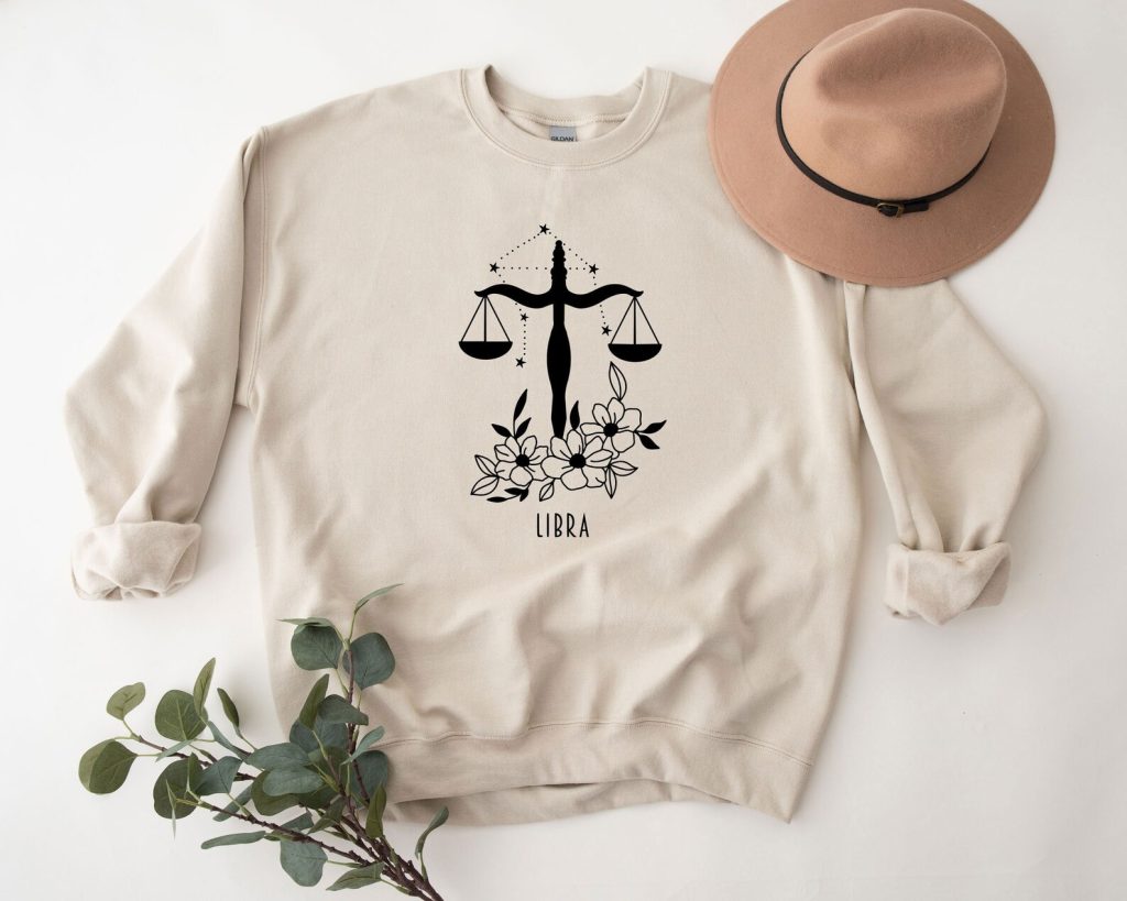 A sand colored crewneck sweatshirt featuring a floral Libra scales constellation graphic, styled with a brown hat and eucalyptus, among cozy October birthday gift ideas.