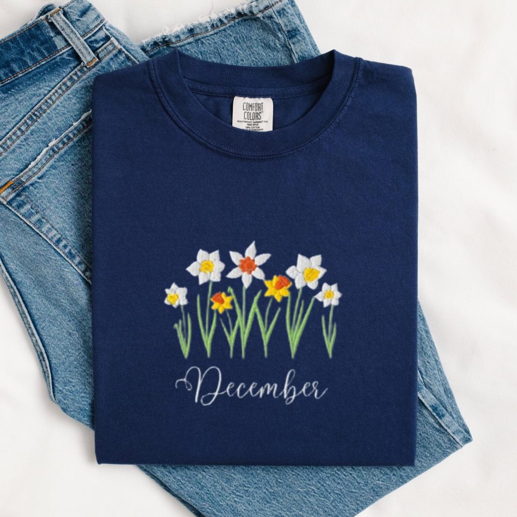 A navy blue t-shirt with embroidered narcissus flowers and the word December, a sweet birth flower December birthday gift idea.