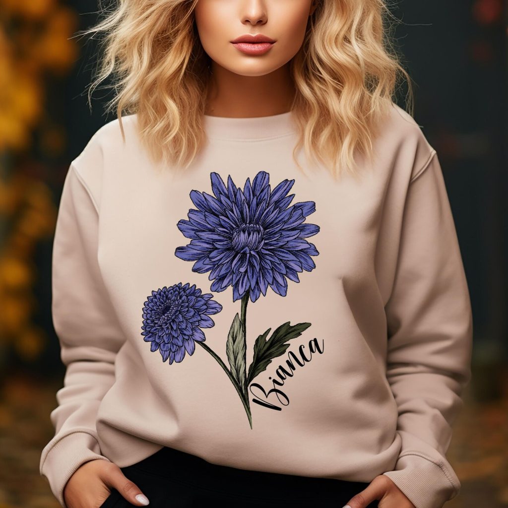 A woman wearing a sand crewneck sweatshirt printed with a bold purple chrysanthemum illustration and personalized name, a cozy November birthday gift idea.
