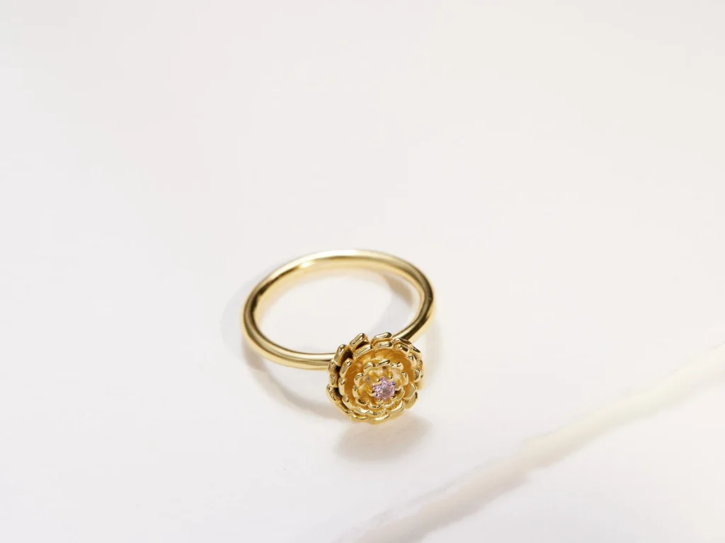 A gold marigold flower ring with a small pink center gemstone on a thin band, one of the most delicate October birthday gift ideas.