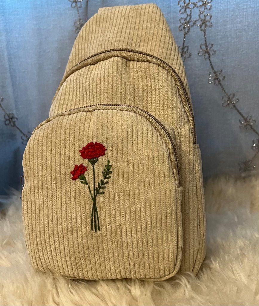 A cream corduroy crossbody sling bag with hand embroidered red carnation birth flowers, a stylish January birthday gift idea.