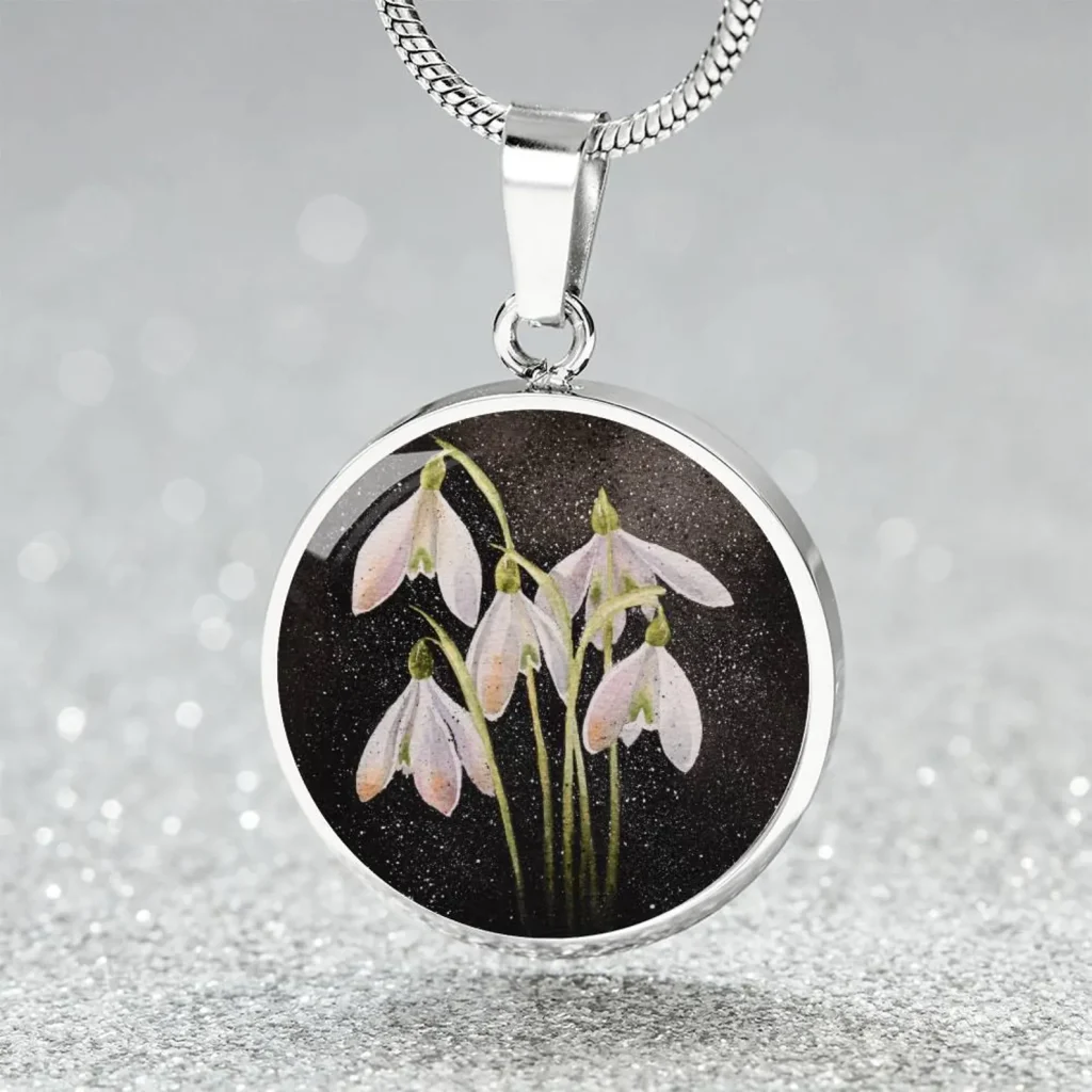 A silver circle pendant necklace featuring a painted snowdrop birth flower design, a pretty and delicate January birthday gift idea.