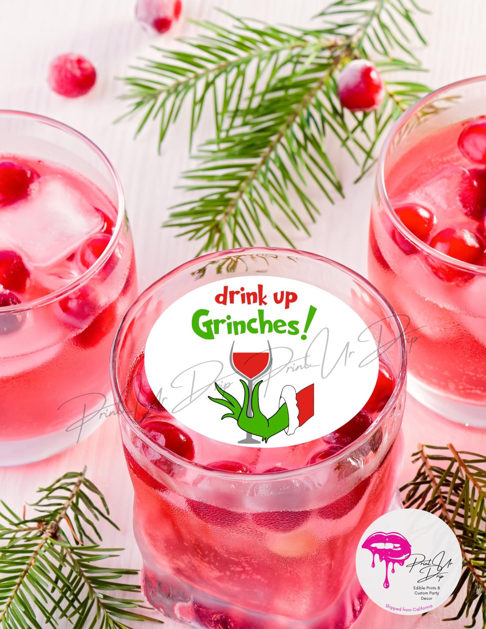 Drink Up Grinches edible drink topper on a cranberry cocktail glass surrounded by festive pine and fresh cranberries, a clever and cheeky Grinch party drinks table detail.
