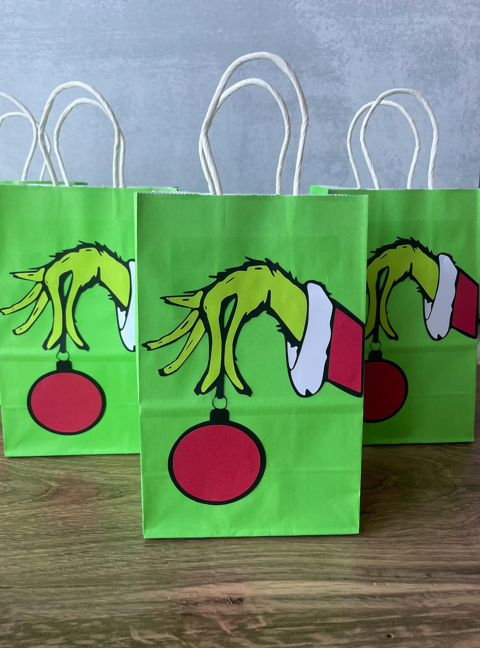 Bright green Grinch hand holding a red ornament printed party favor bags with white rope handles, charming and festive Grinch party goodie bags guests will love taking home.