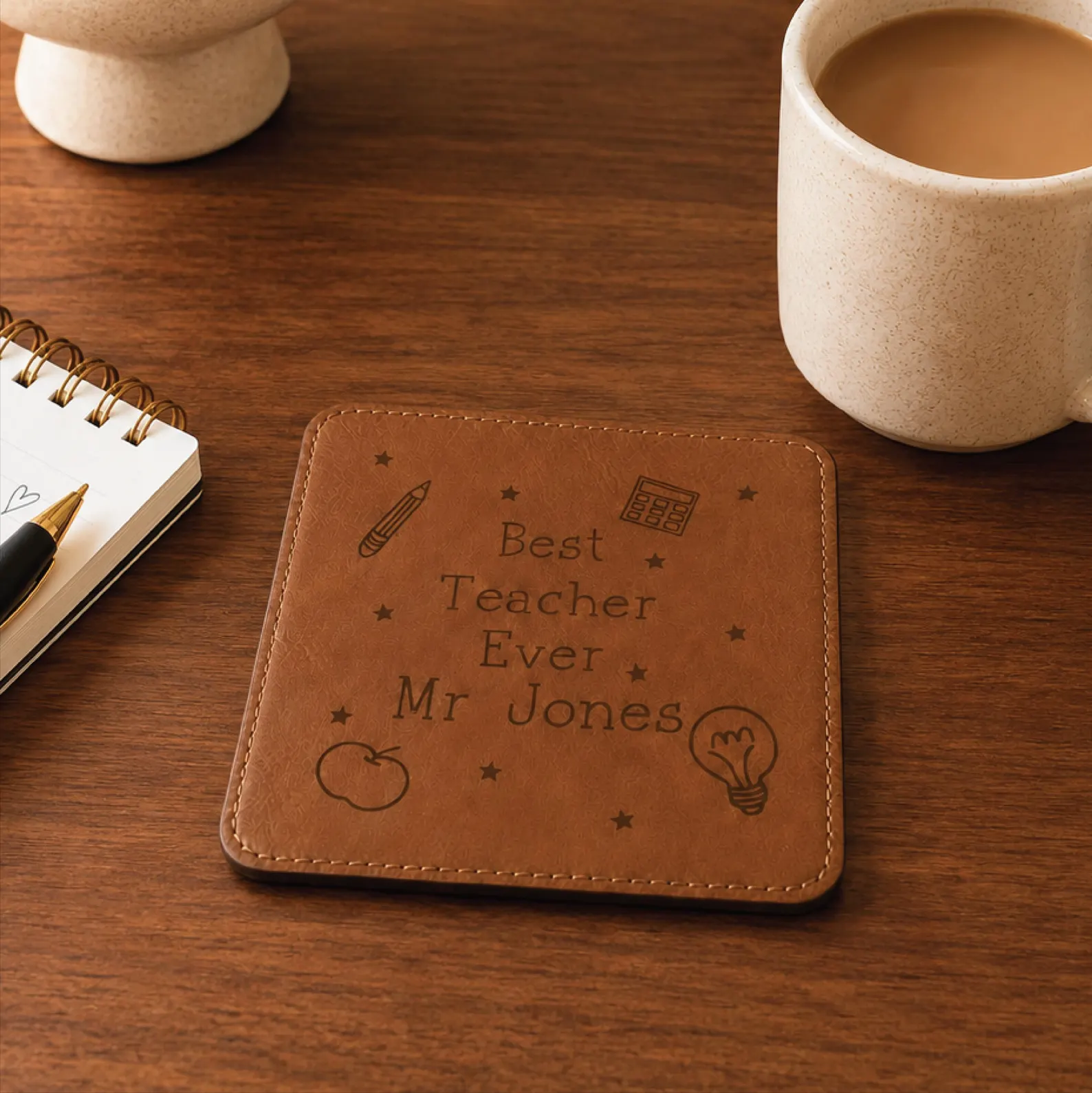 Personalized engraved tan leather coaster with Best Teacher Ever name and school icons on a wood desk beside a coffee mug, a simple yet stylish male teacher appreciation gift ideas he will use every day.
