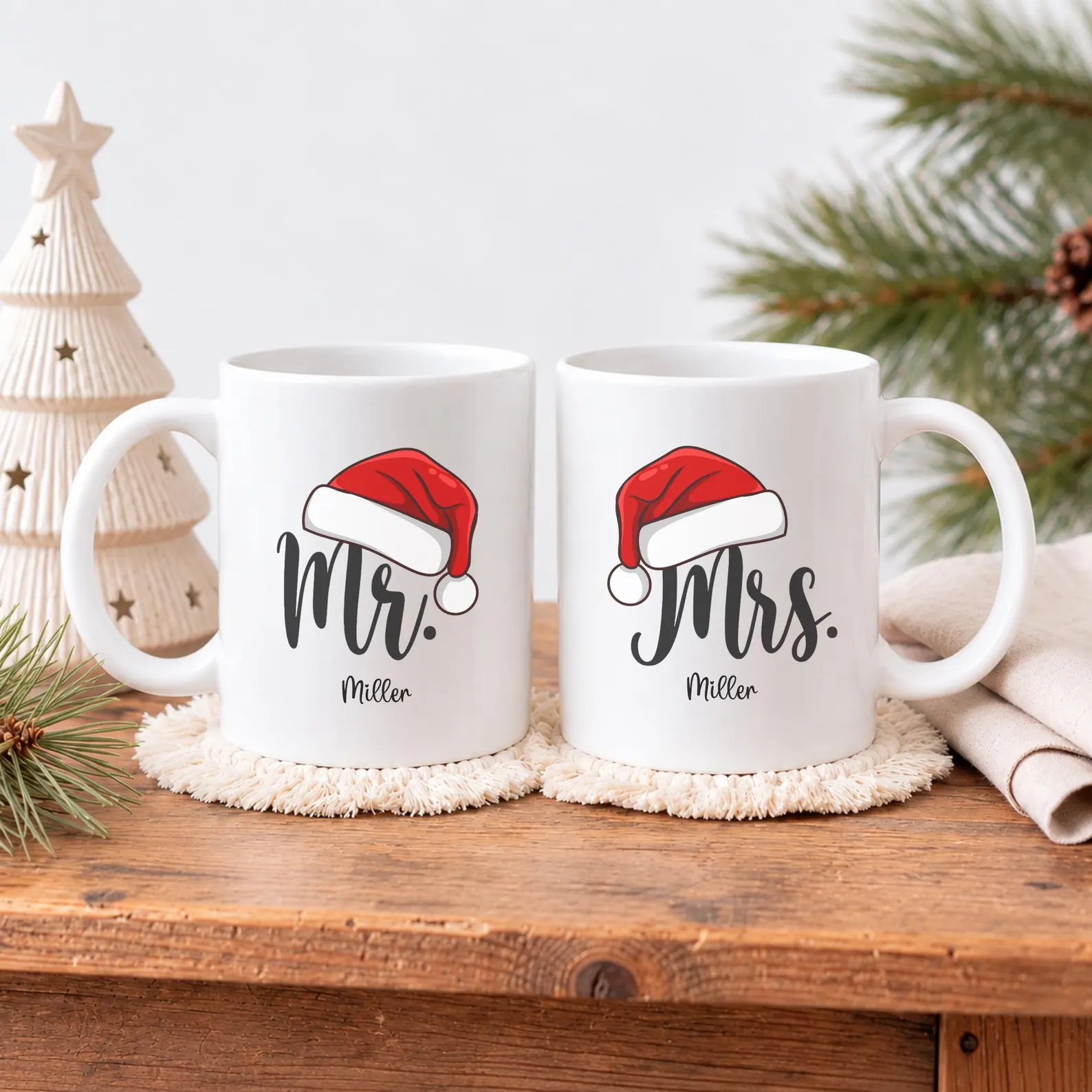 Personalized Mr and Mrs Santa hat couples mug set with family name on a rustic wood surface beside a ceramic Christmas tree, a sweet and thoughtful Christmas hostess gift ideas for the couple who loves to host.
