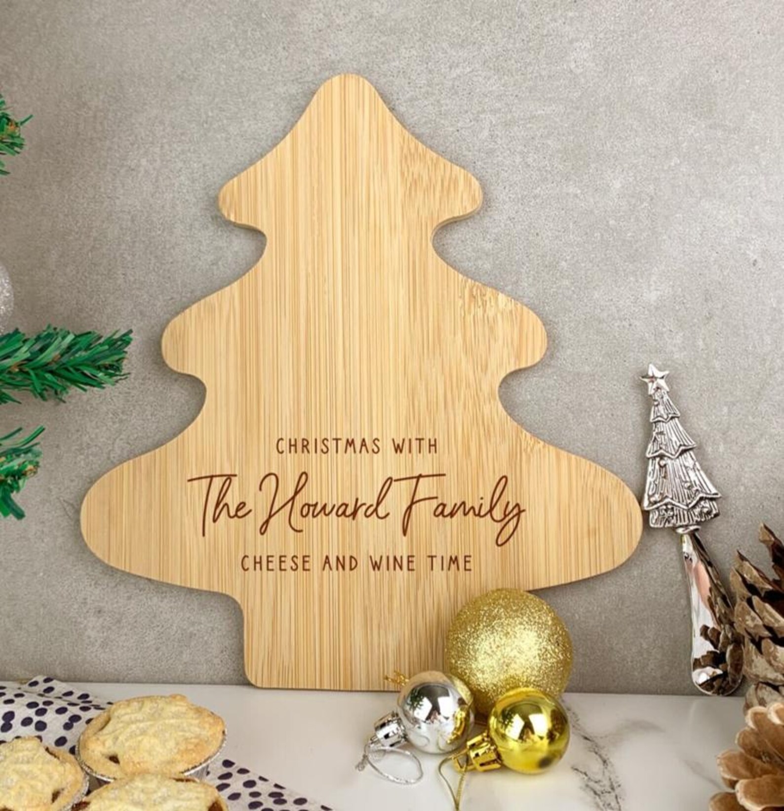 Personalized Christmas tree shaped bamboo cheese and wine board engraved with family name displayed beside gold ornaments and mince pies, a show stopping and keepsake worthy Christmas hostess gift ideas for entertaining.