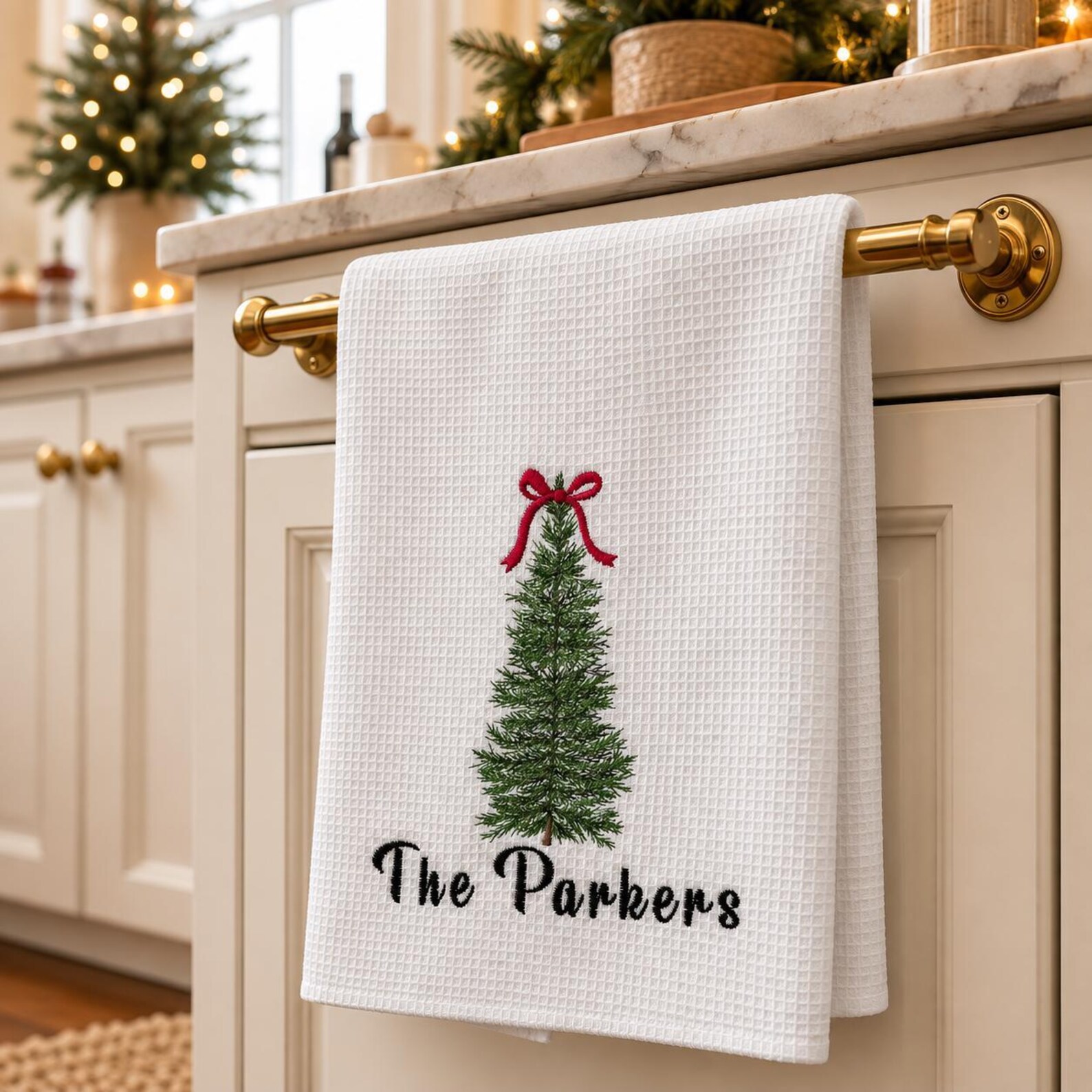Personalized white waffle weave kitchen towel embroidered with a Christmas tree and red bow topped with a family name hanging on a brass rail in a beautifully styled kitchen, a sweet and practical Christmas hostess gift ideas she will display all season long.