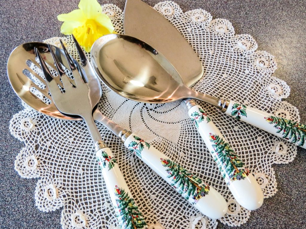 Stainless steel serving set with holly and Christmas tree patterned ceramic handles including salad fork, spoon and cake server on a lace doily, a timeless and elegant Christmas hostess gift ideas for the one who loves to entertain in style.